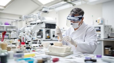Mexican lab technician hand-coloring all-porcelain bridge with CAD/CAM machines behind